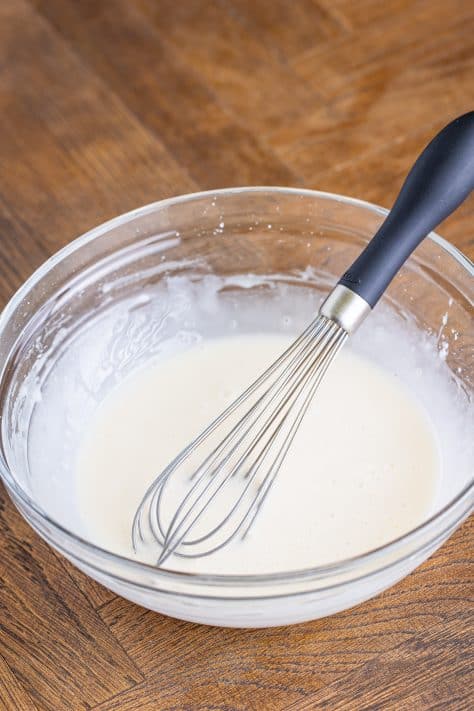 A whisk in a bowl of powdered sugar, milk, and vanilla mixed together.