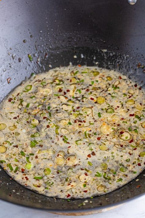 A skillet with green onions, red pepper flakes, garlic, oil, chicken stock, honey, soy sauce, cornstarch slurry, and rice wine vinegar.