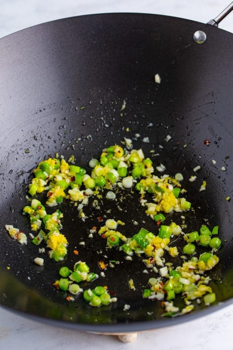 A wok with green onion, garlic, ginger, and chili flakes.