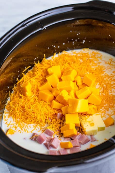 A Crock pot with ham, macaroni, milk, evaporated milk, 3 cups of shredded cheddar cheese, Velveeta, butter, salt, pepper, and ground mustard in it.