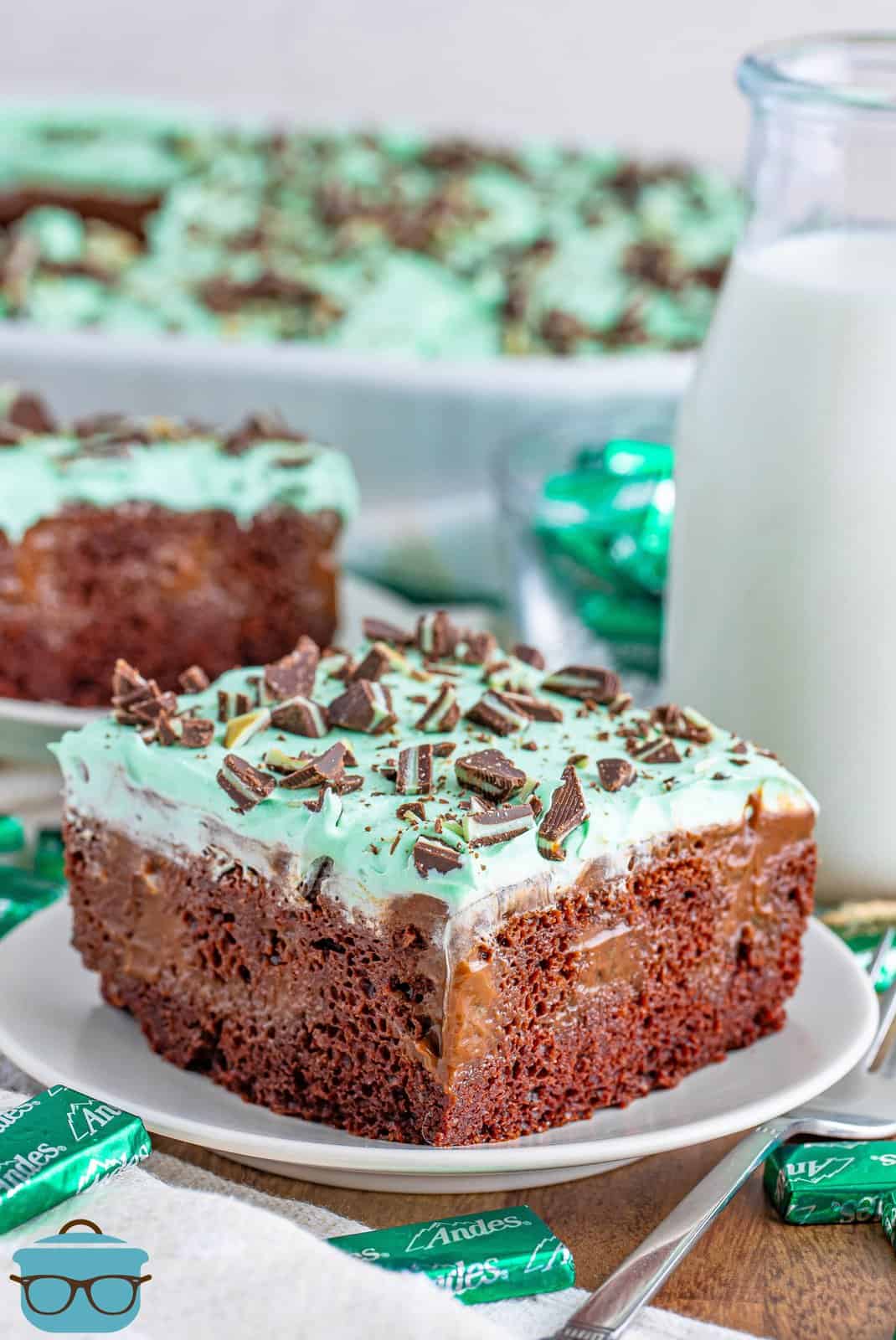 A piece of homemade Mint Chocolate Poke Cake on a white serving plate in front of the rest of the cake in a baking dish.