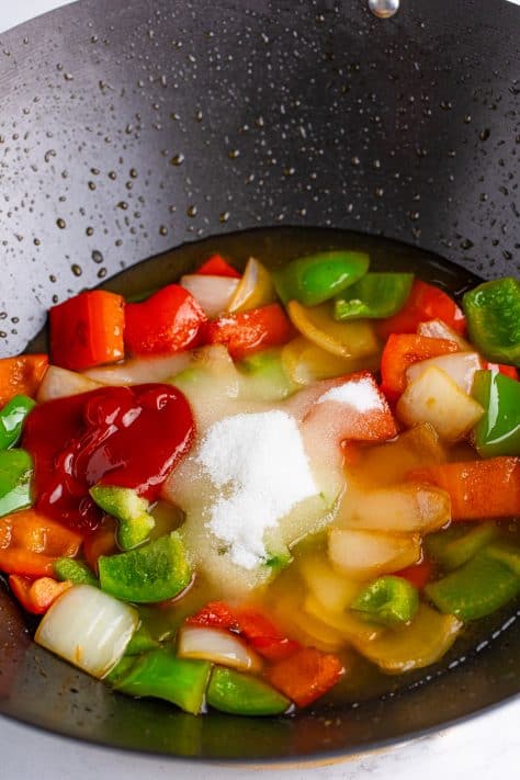 A wok with green bell pepper, red bell pepper, diced onion, water, pineapple juice, vinegar, sugar, ketchup, and salt.