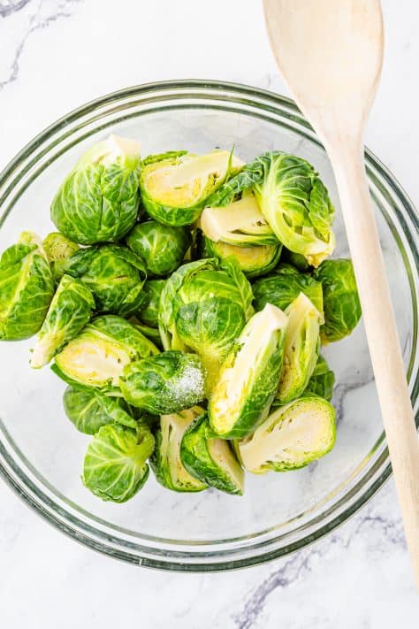 A glass mixing bowl with brussels sprouts, oil and salt.