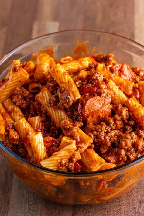 Cooked rigatoni, pizza sauce, pepperoni, sausage, ground beef, and onion in a bowl.