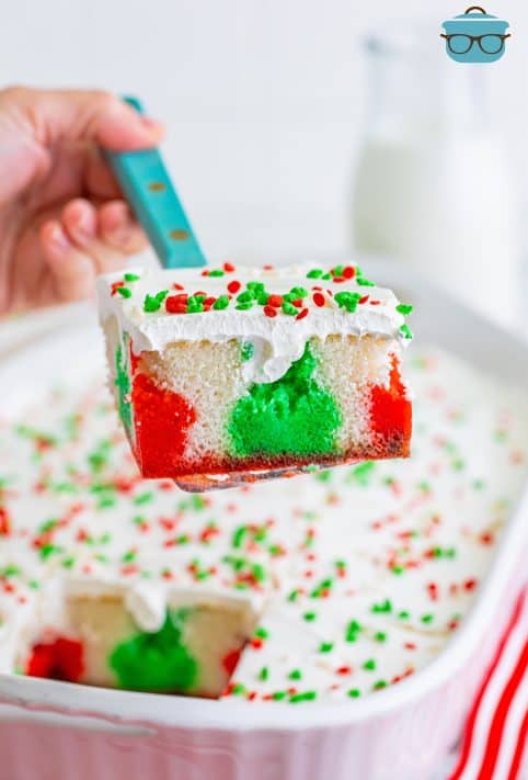 A serving utensil holding a slice of Christmas Jello Poke Cake above the rest of the cake.