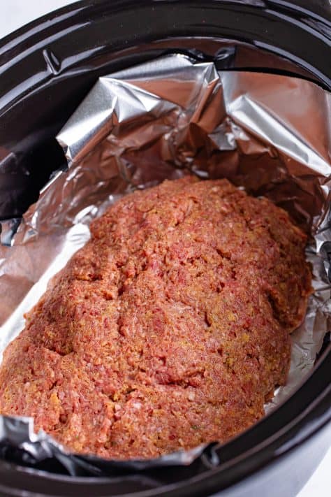 An oval meatloaf mixture in a slow cooker lined with aluminum foil.