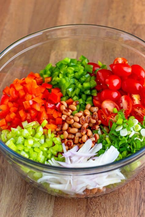 A glass bowl of black-eyed peas, tomatoes, red bell pepper, green bell pepper, celery, green onions, and shallots.