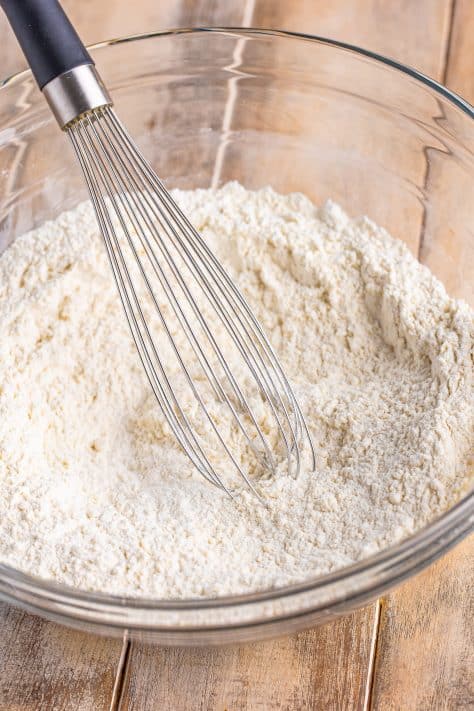 A whisk, flour, baking powder, and sugar in a glass bowl.
