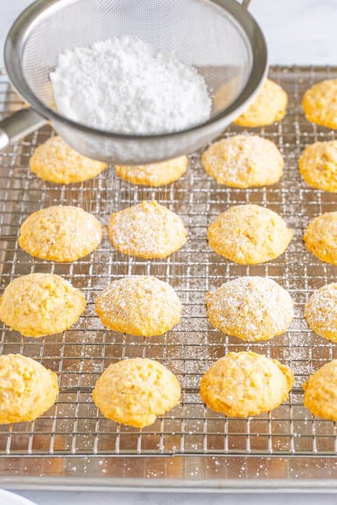 Powdered sugar being sifted through a sifter on top of cookies on a wire rack.