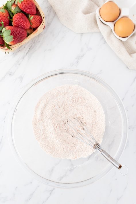 A glass mixing bowl with flour, baking powder, salt, and strawberry jello powder.