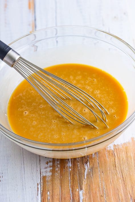 A glass mixing bowl with caramel sauce and sweetened condensed milk mixed with a whisk.