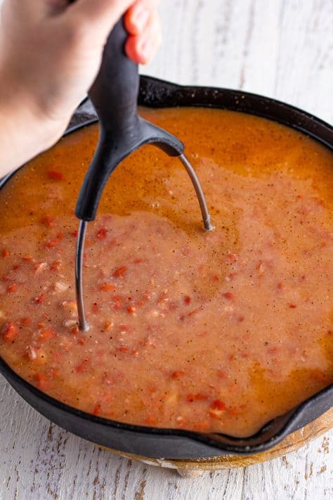 A potato masher mashing red beans and seasonings with water in a skillet.