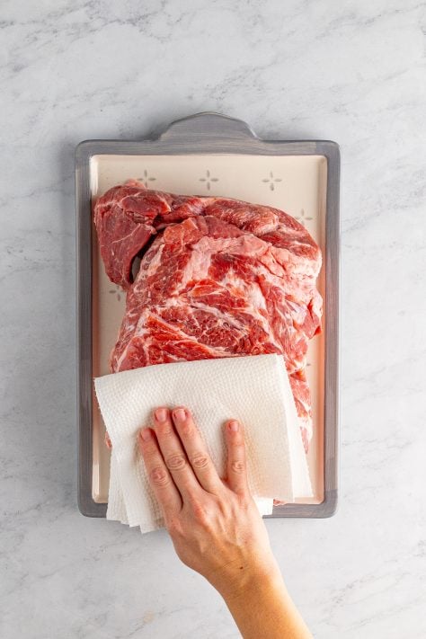 A hand holding a paper towel drying off a pork shoulder.