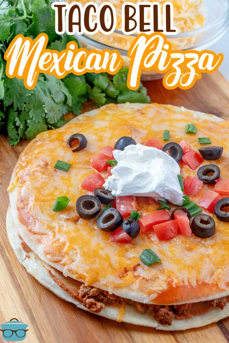 Fully baked Mexican Pizza shown on a wooden cutting board and topped with diced tomatoes and sliced black olives.