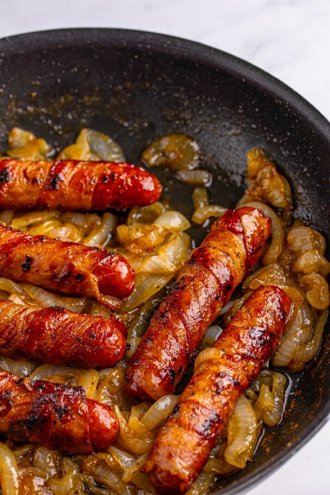 Bacon wrapped brats in a bunch of cooked onions in a skillet.