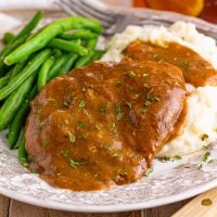 Cubed Steak shown covered in gravy on a plate with green beans and mashed potatoes.