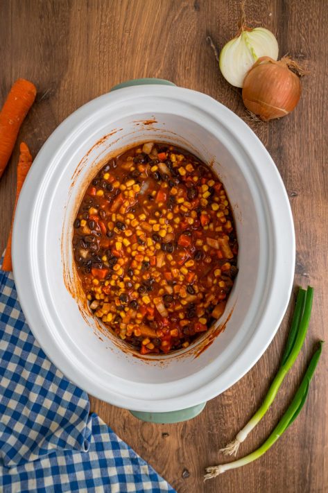 A Slow Cooker with chicken breast, diced onion, diced carrot, black beans, corn, enchilada sauce, tomato sauce, and taco seasoning.