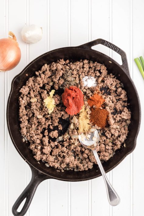 Tomato paste, garlic, and seasonings being added to ground beef and onions in a skillet.
