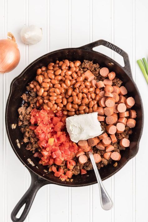 A ground beef, onion and seasonings skillet with hotdog slices, Rotel, chili beans, and cream cheese being added.