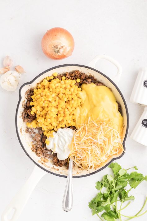 A skillet with ground beef, onion cheese, corn, cream of chicken, sour cream, and milk.
