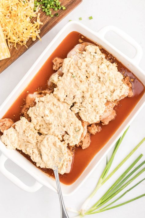 Cream cheese mixture on taco seasoned chicken with refried beans and enchilada sauce in a baking dish.