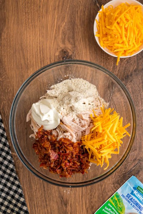 A bowl with shredded chicken, sour cream, ranch seasoning, shredded cheddar cheese, and crumbled bacon.