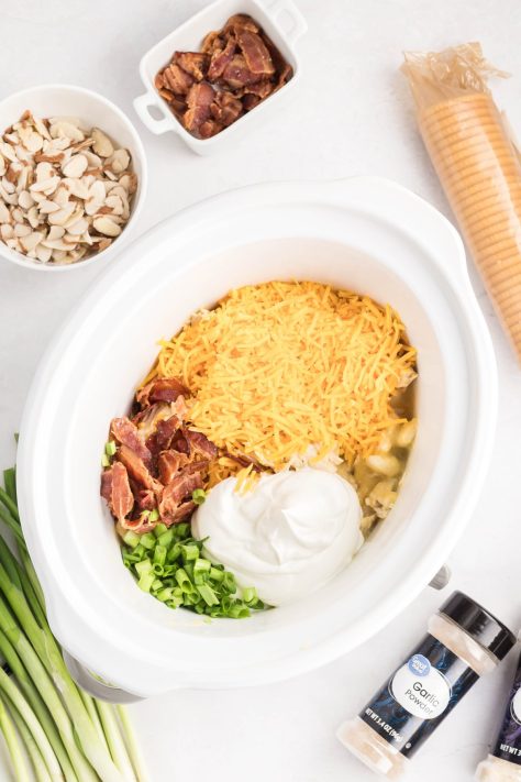 A Slow Cooker with a chicken mixture, cheddar cheese, sour cream, half of the bacon, and green onions.