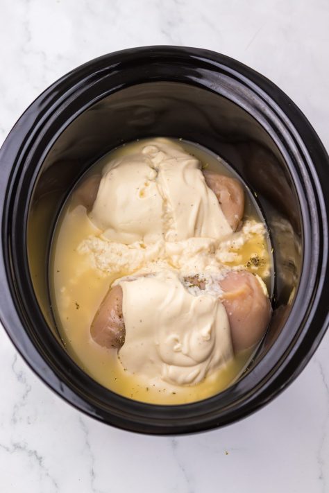 A Slow Cooker with chicken breasts, alfredo sauce, chicken broth, garlic, Italian seasoning, and pepper.