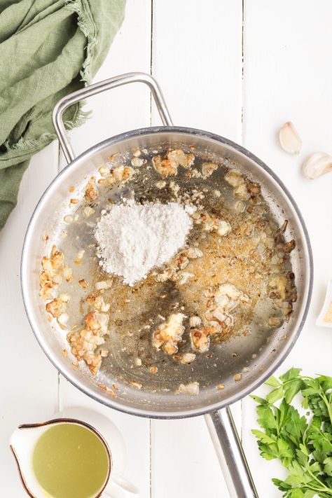 Flour being added to a skillet with butter, shallots, and garlic.