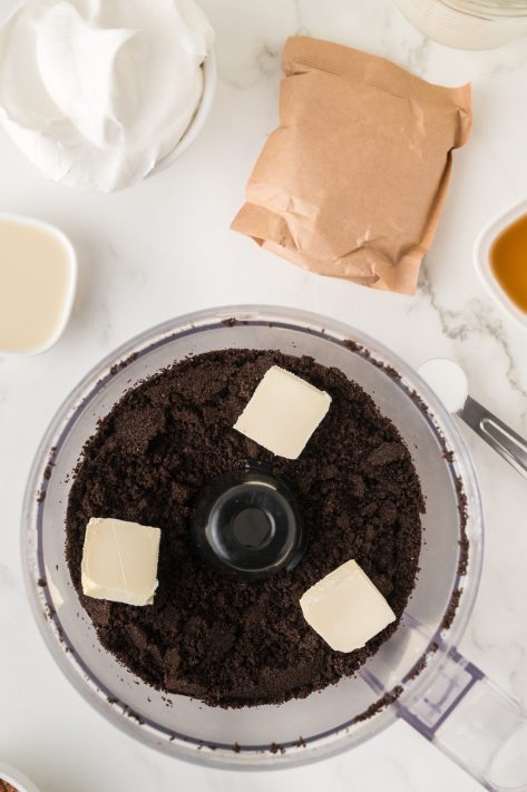 Pads of butter being added to Oreo crumbs in a food processor.