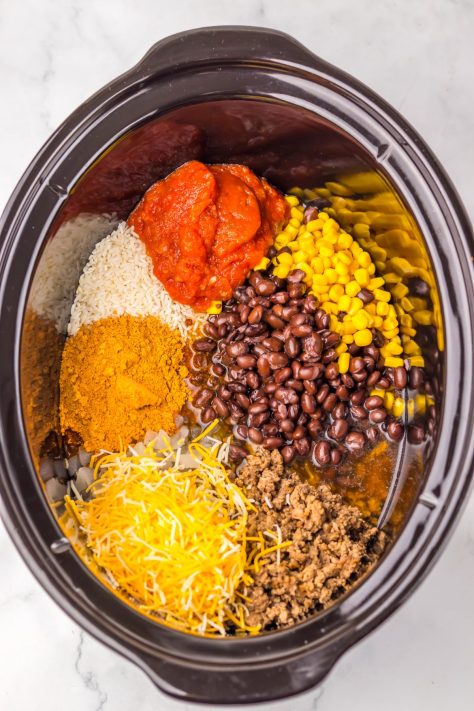 A Slow Cooker with cooked beef and onion, taco seasoning, uncooked rice, beef broth, black beans, corn, and salsa and 1 cup shredded Mexican cheese blend.