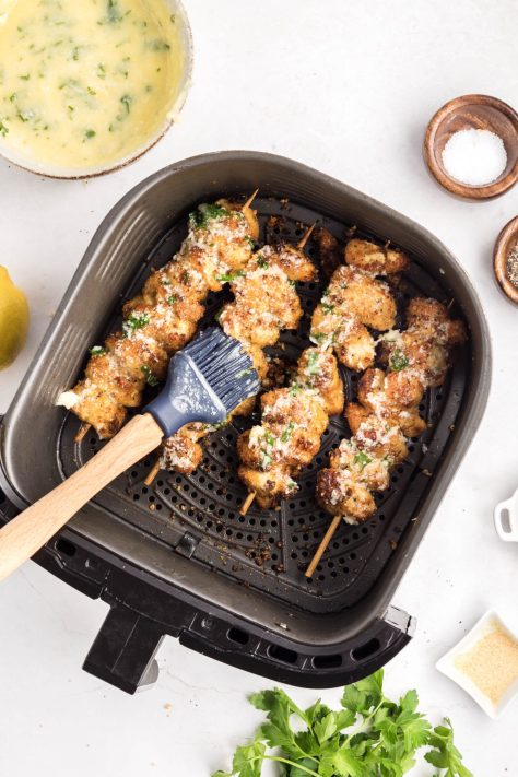 A pastry brush brushing the lemon garlic butter sauce over the chicken skewers in an Air Fryer basket.