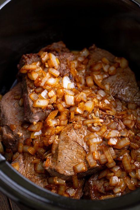 Onions and garlic being added to seared steaks in a Crock Pot.