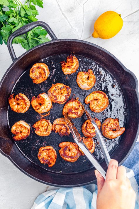Shrimp being cooked in a skillet and a pair of tongs getting ready to flip them over.