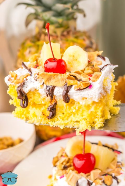 A slice of Banana Split Poke Cake being held above the rest of the cake.