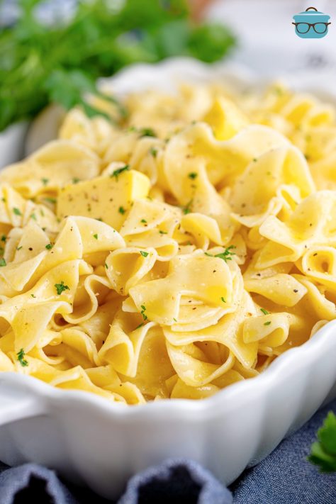 A baking dish with Buttered Noodles and a serving spoon stirring them.