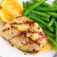 Looking closely at a piece of Grilled Sea Bass with sauce, green beans, and a lemon wedge.